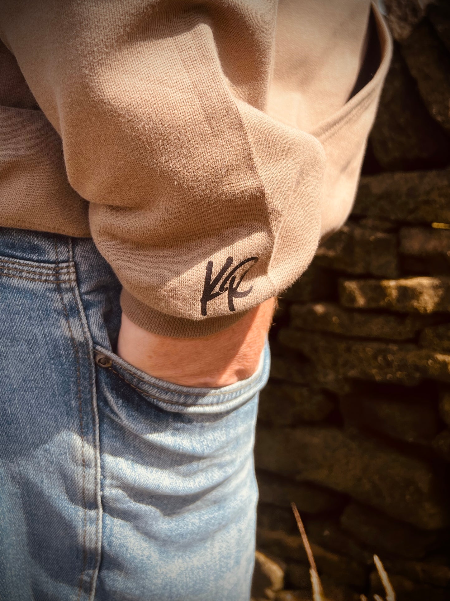 Close-up of a beige sleeve with a logo, worn over blue jeans against a stone wall background.