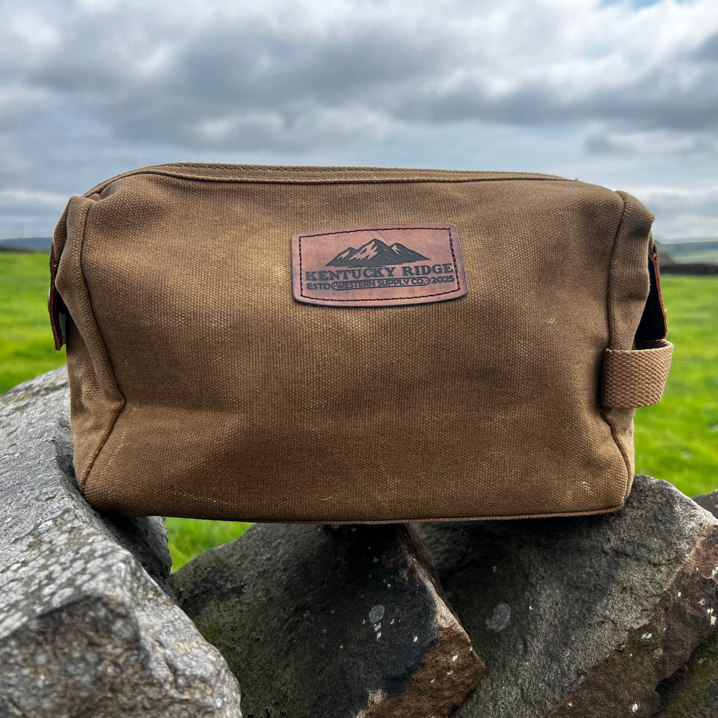 Brown washbag with Kentucky Ridge logo on a stone wall against a green field and cloudy sky